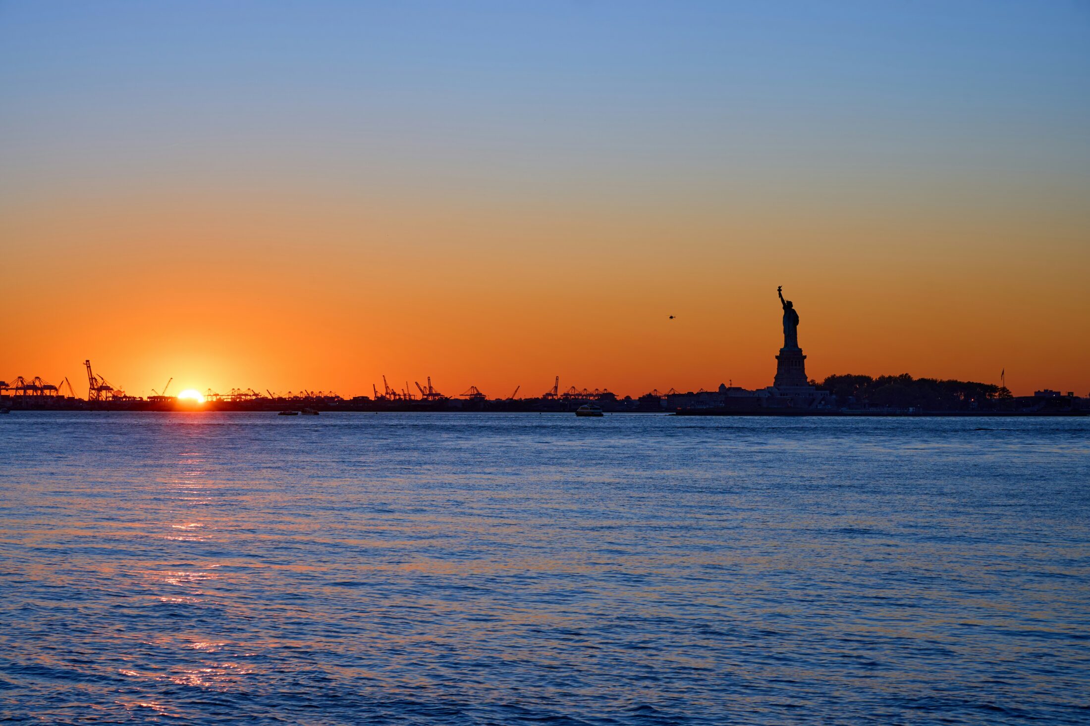 The sun is setting over a body of water and a silhouette of the Statue of Liberty