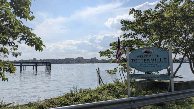 Welcome to Tottenville sign at the shore with the fishing pier and Raritan Bay behind it
